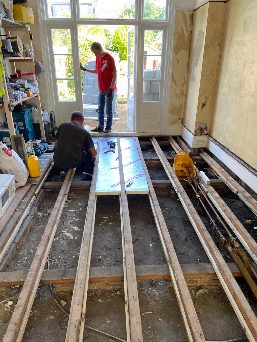 Two workers laying insulation panels between exposed joists ahead of Flooring Installation during home renovation