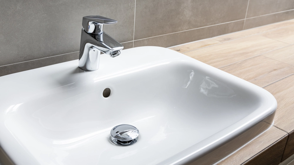 White ceramic sink with chrome faucet and pop-up drain installed on wood countertop against tiled wall, showcasing Bathroom Fitting