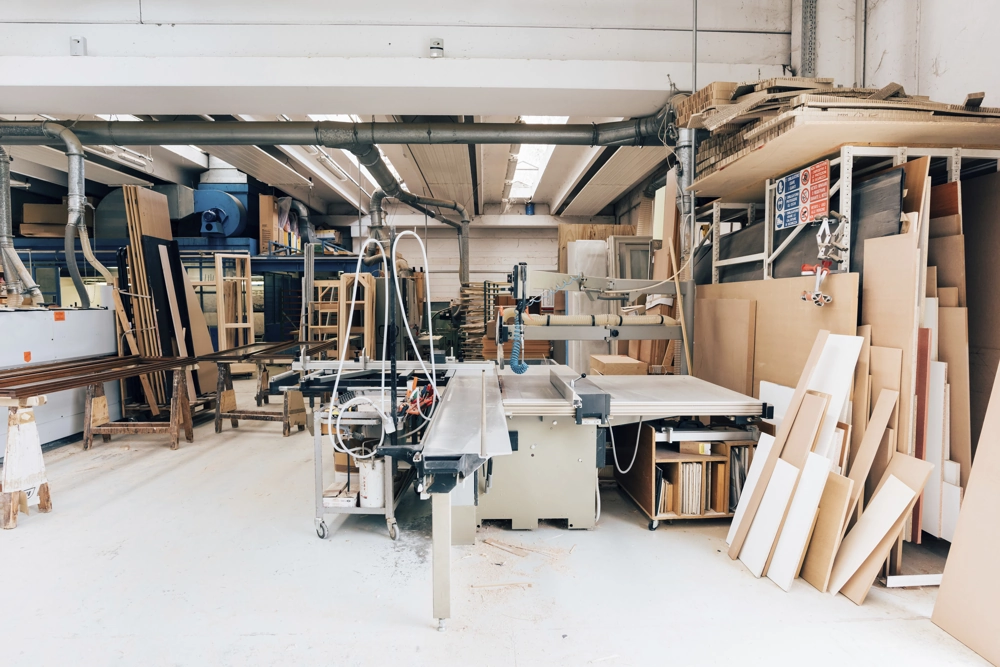 Carpentry workshop with industrial table saw, dust extraction pipes and stacked wood panels ready for bespoke furniture
